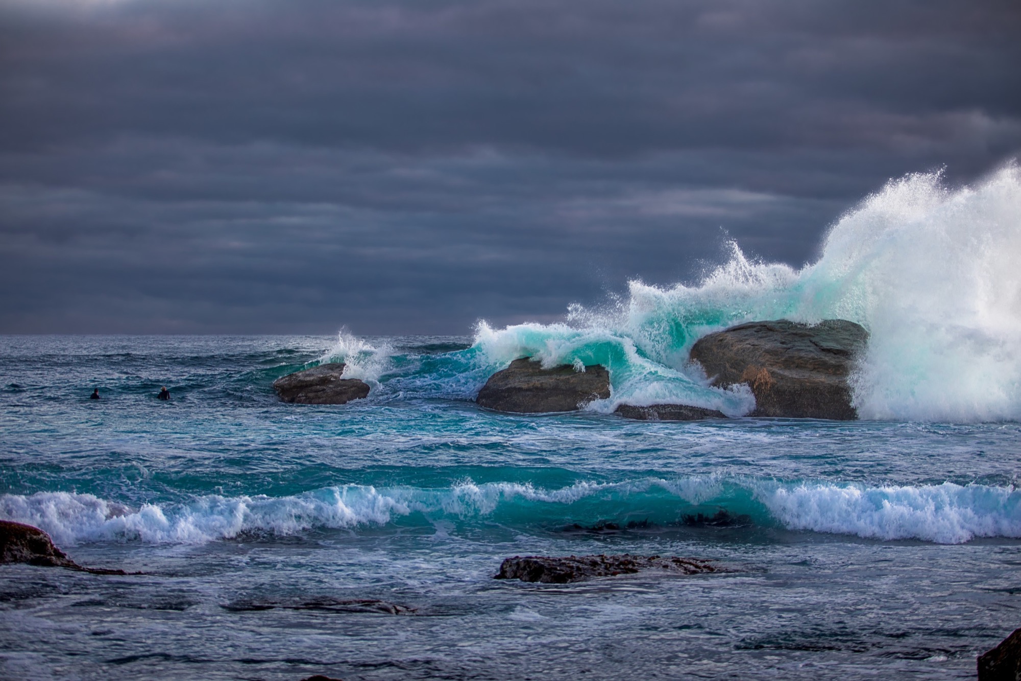 A winter wave breaking over dark coastal rocks, with white spray and a heavy sky