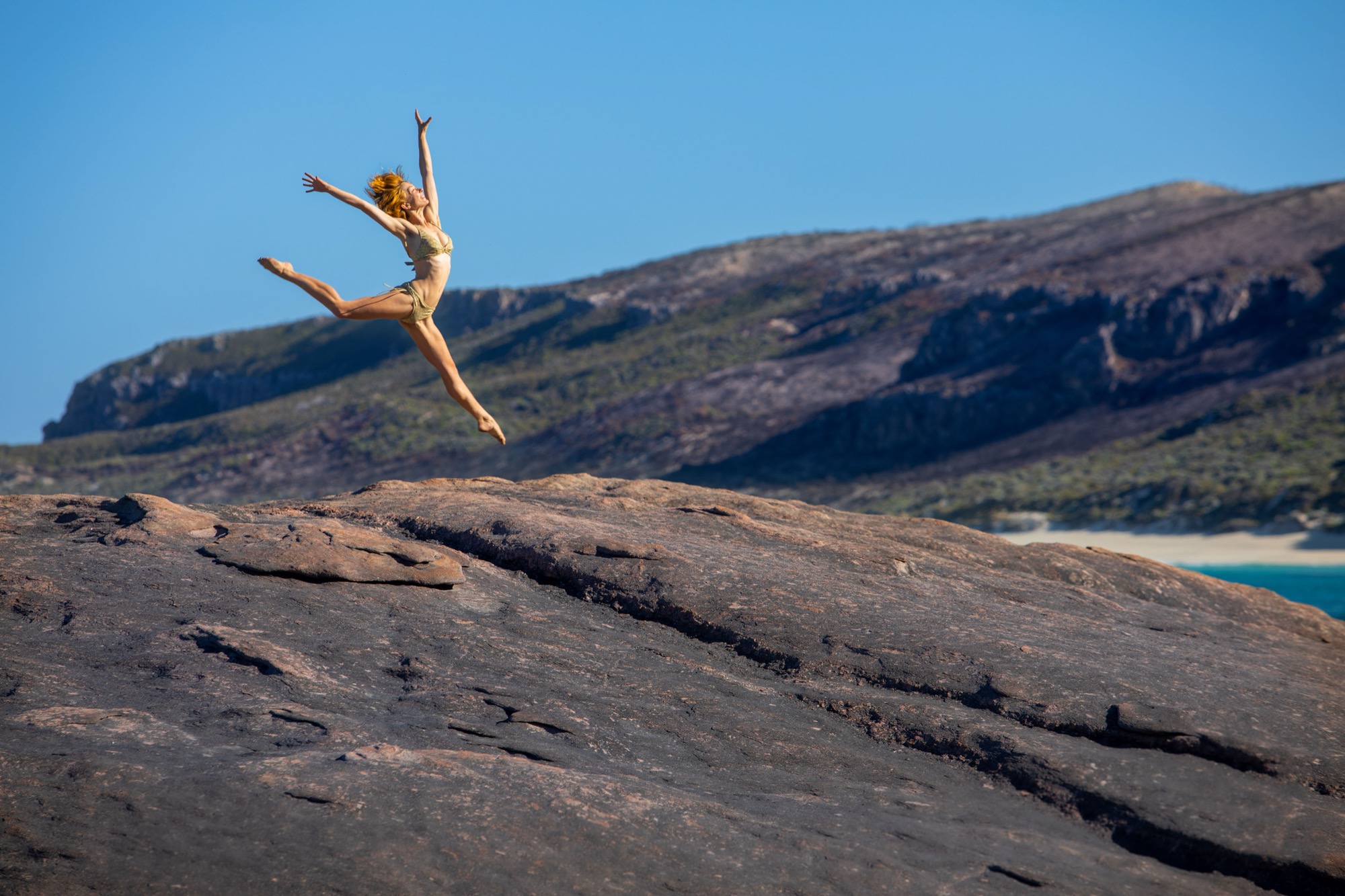 A woman leaping above a coastal rock at Contos, with scrub-covered hills and a strip of beach behind her