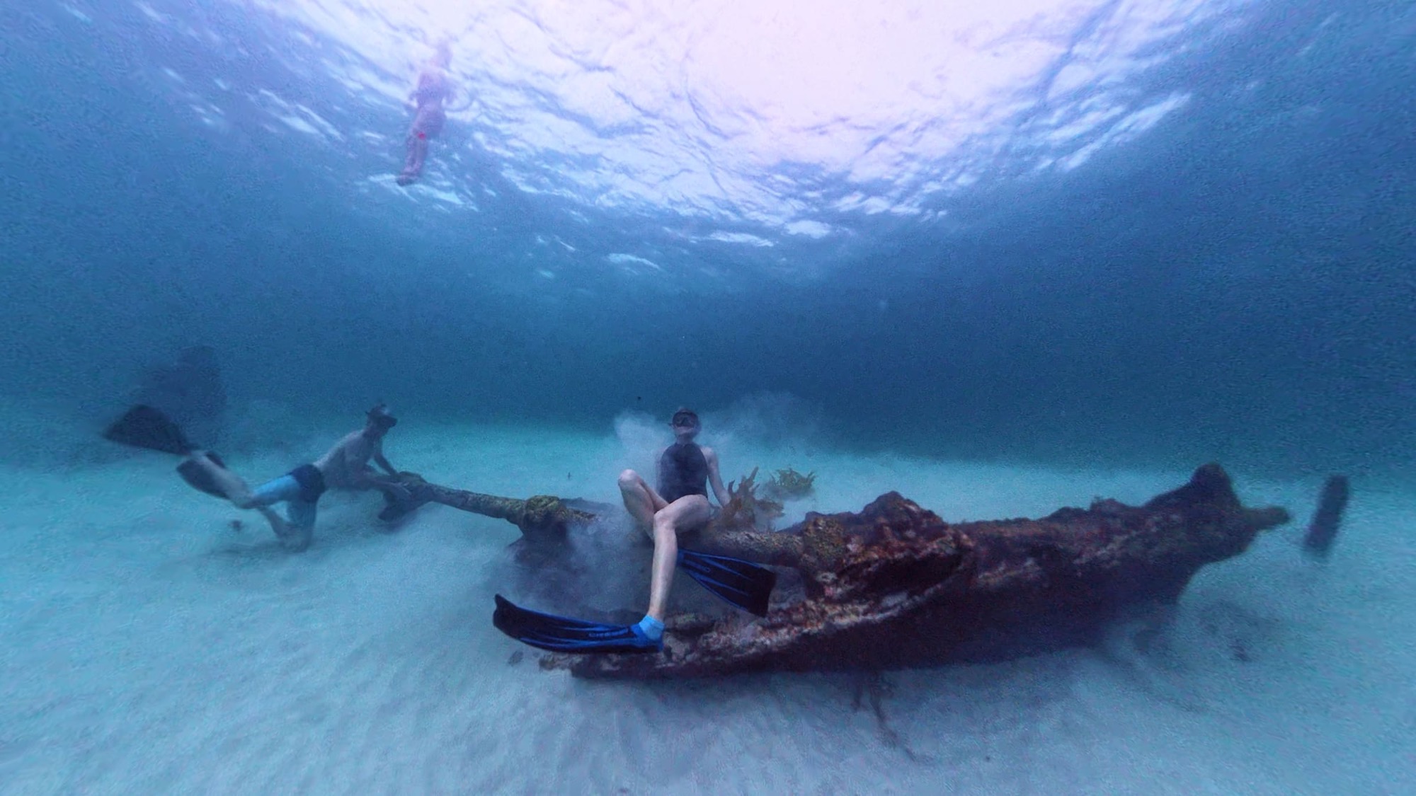Three swimmers exploring a rusted shipwreck on a pale sandy seabed in clear blue-green water